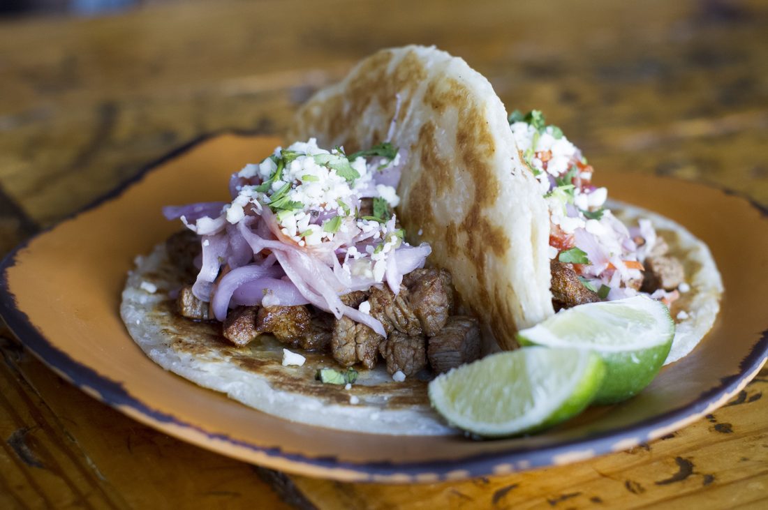 A paratha taco on a wooden plate, filled with spiced ground lamb, pickled red onion and herbed crema, with lime wedges alongside.