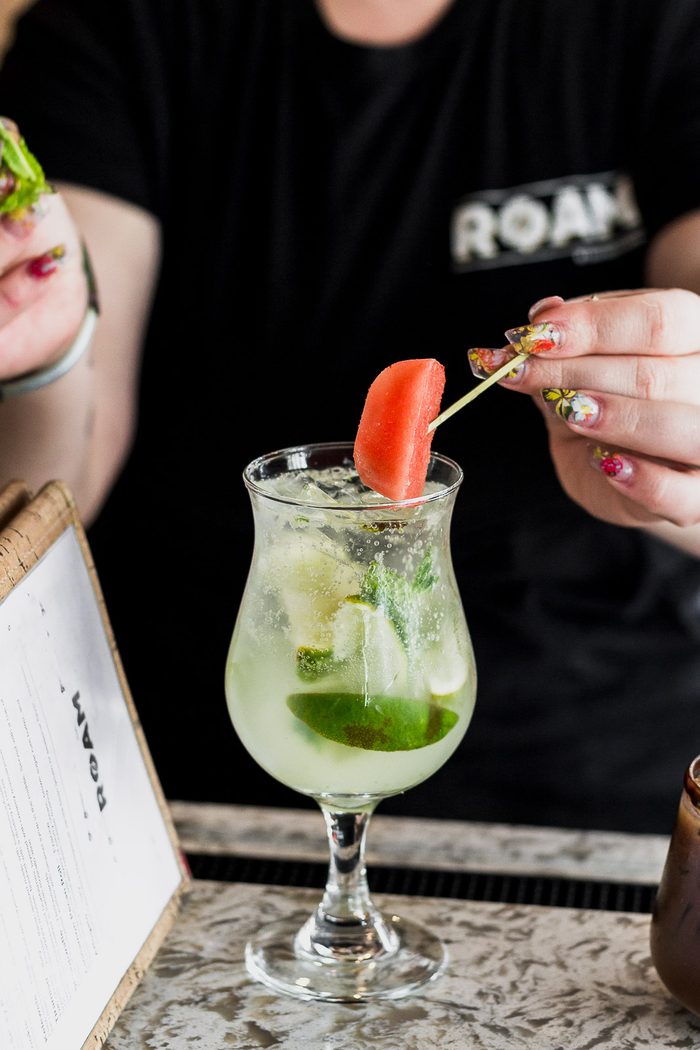 A server in a ROAM shirt setting down a tall cocktail garnished with mint and watermelon at a marble table.